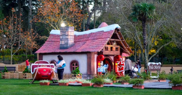 Pátio com a casa do Papai Noel, enfeitada com luzes de Natal. Uma duende recebe os convidados na porta da casa. trenó vermelho do Papai Noel ao lado da casa